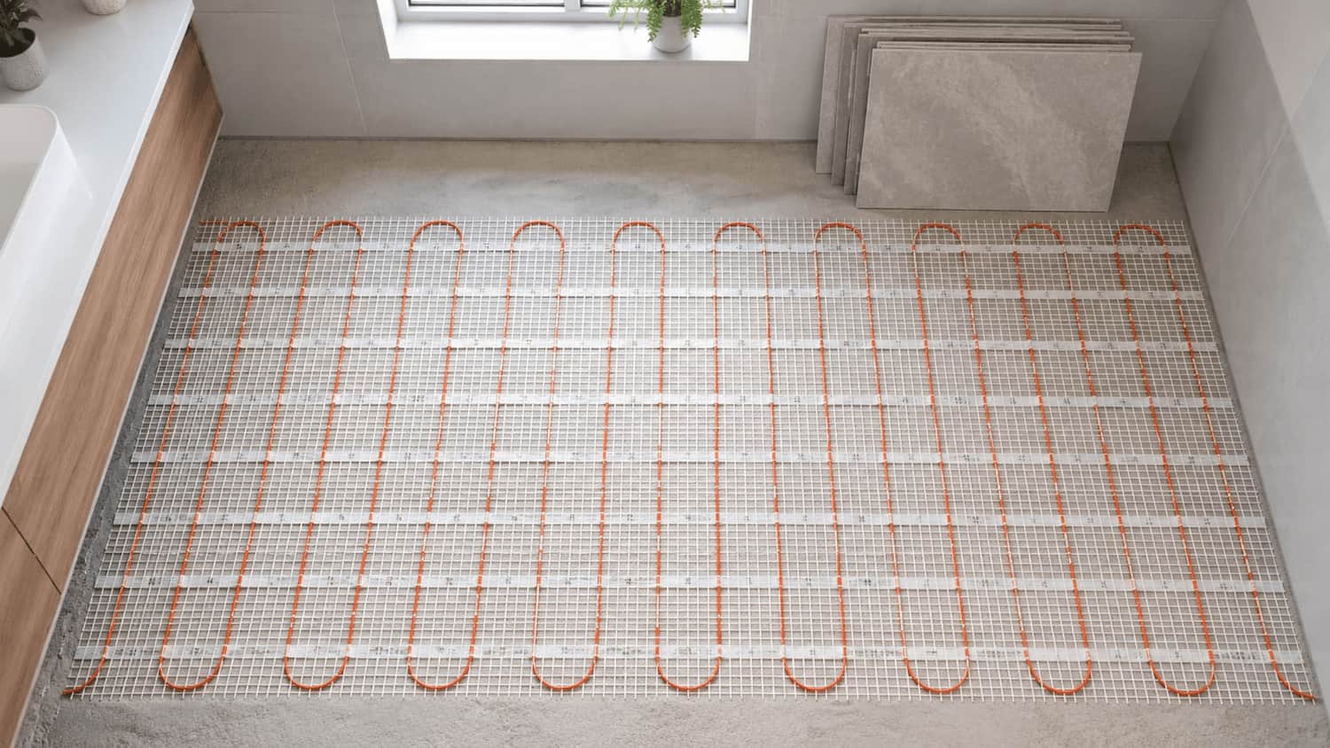 Electric underfloor heating mat with orange resistance cable laid on concrete subfloor, ceramic tiles stacked at the edge